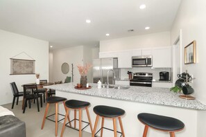 Een moderne keuken met witte kasten, een granieten aanrechtblad en roestvrijstalen apparatuur. Er staan drie barkrukken en een eettafel met stoelen.
