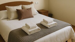 A neatly made bed with white and brown bedding, a painting of autumn trees, an air conditioning unit, and a window with wooden shutters.