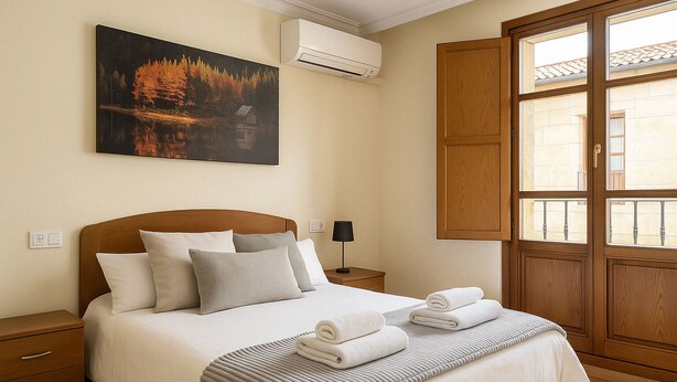 A neatly made bed with pillows and a folded towel, a bedside lamp, a painting of a forest scene, and a window with wooden shutters.