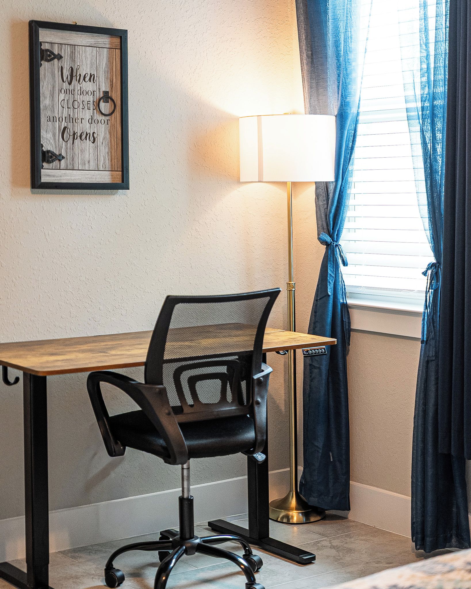 Habitació d'hotel amb escriptori, una cadira, una làmpada i una finestra amb cortines.