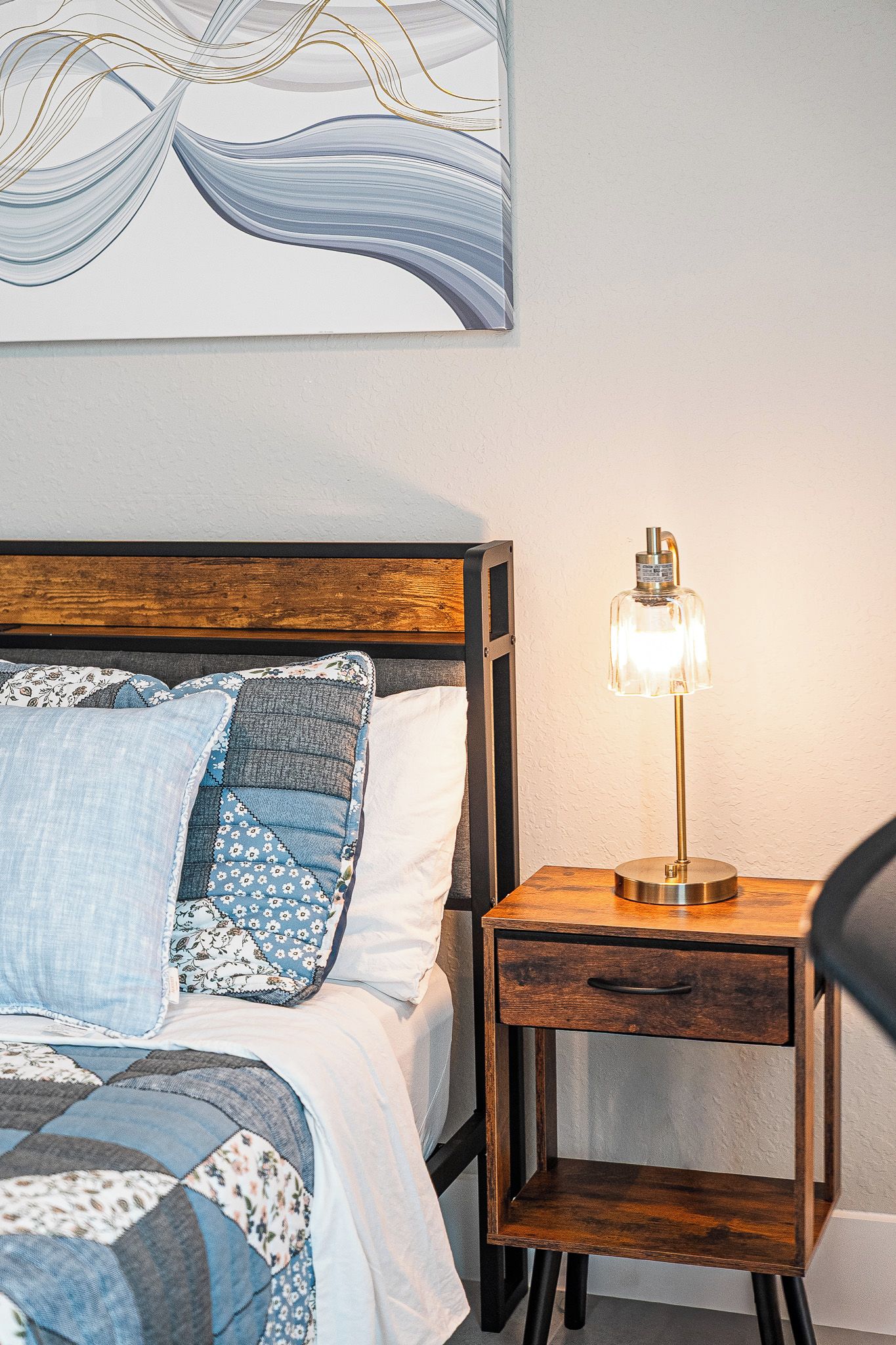 A neatly made bed with a wooden headboard, a bedside table with a lamp, and a wall with a piece of abstract art.