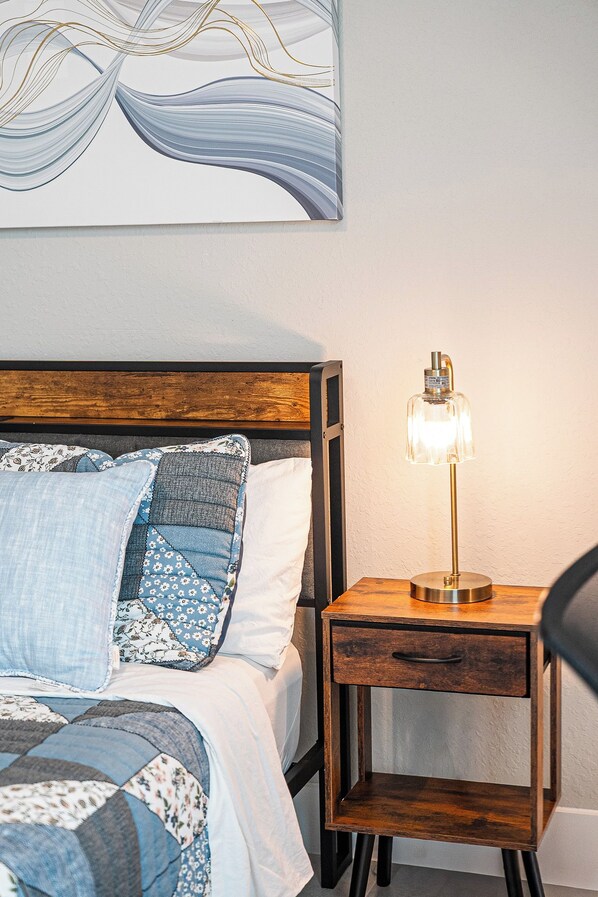 A neatly made bed with a wooden headboard, a bedside table with a lamp, and a wall with a piece of abstract art.