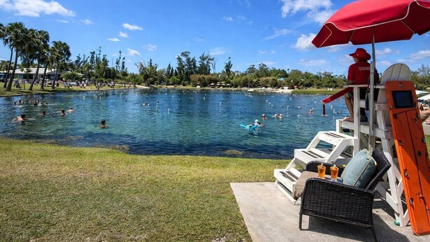 A pool area with people swimming and lounging, a lifeguard stand with a red umbrella, and a view of the water and surrounding greenery.