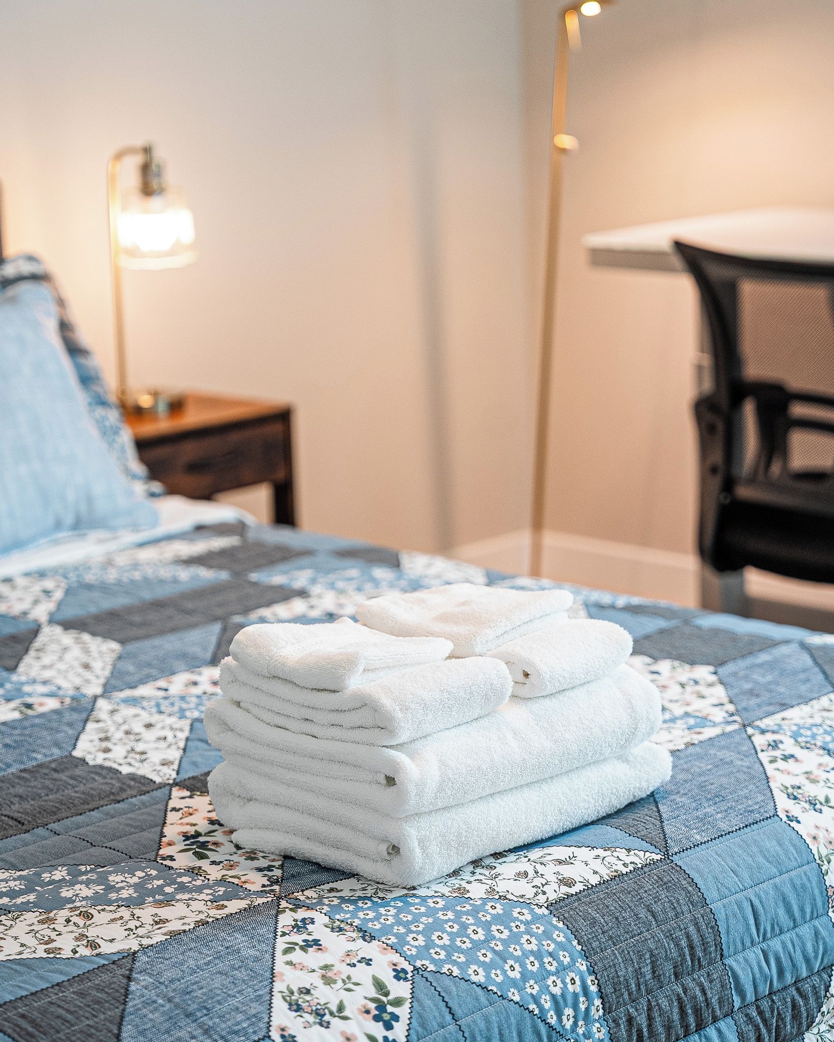 A neatly made bed with a blue floral quilt, a stack of white towels, and a bedside lamp.