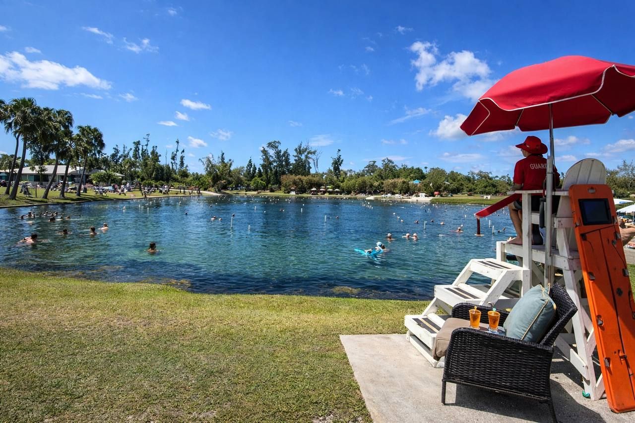 Ein Poolbereich mit Schwimmern und Liegebereich, ein Rettungsschwimmerstand mit rotem Sonnenschirm und Blick auf das Wasser und die grüne Umgebung.