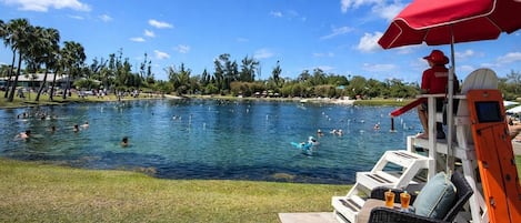 Zona de piscina con gente nadando y descansando, un puesto de socorrista con una sombrilla roja, y vistas al agua y la vegetación circundante.