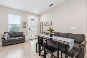 A modern living room with a grey sofa, a dining table with benches, and a kitchen area visible through an open door. - Luxury Healing Retreat North Port (North Port)
