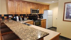 A kitchen with granite countertops, wooden cabinets, a microwave, stove, refrigerator, and a tiled backsplash.