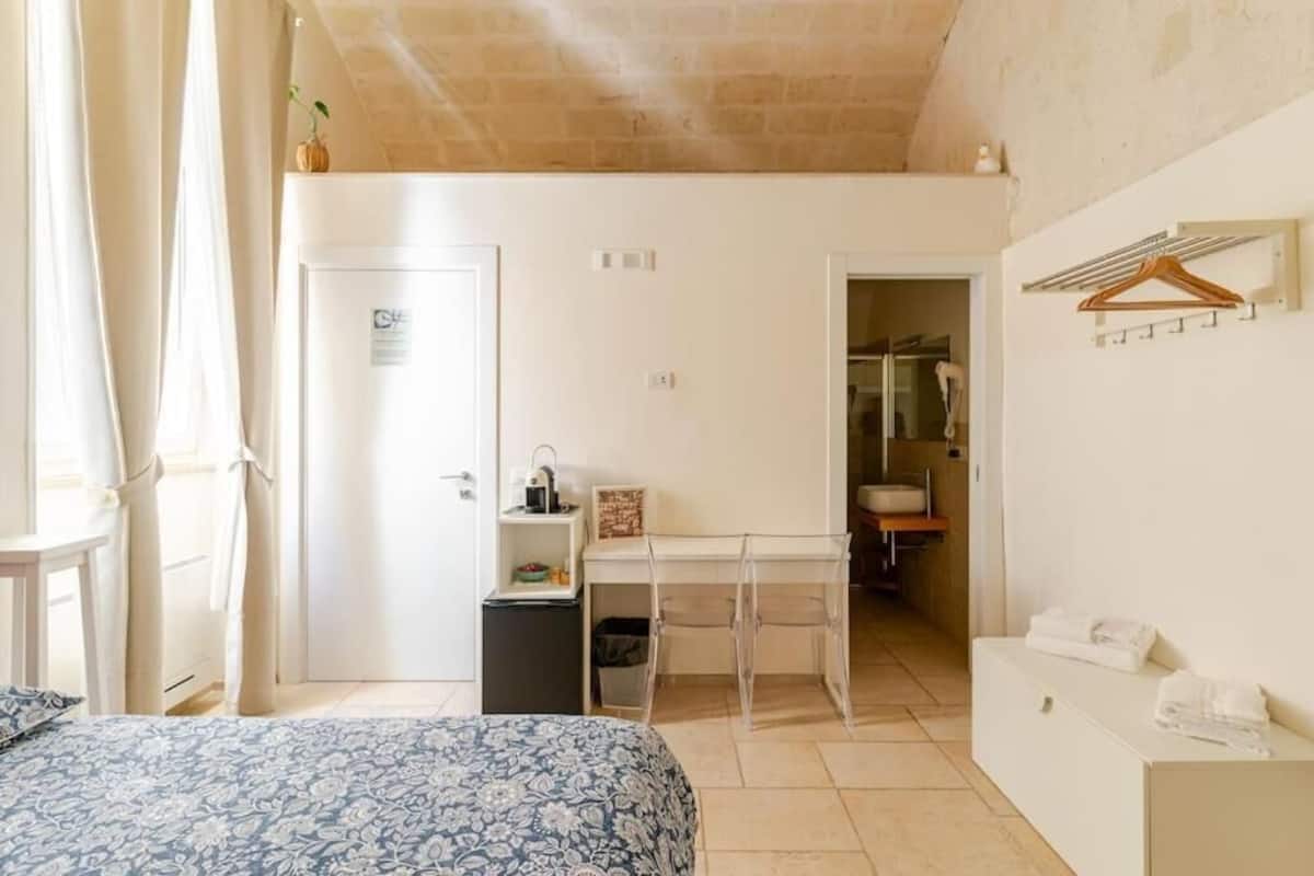 A bedroom with a bed, a desk, a small cabinet, and a bathroom visible through an open door.