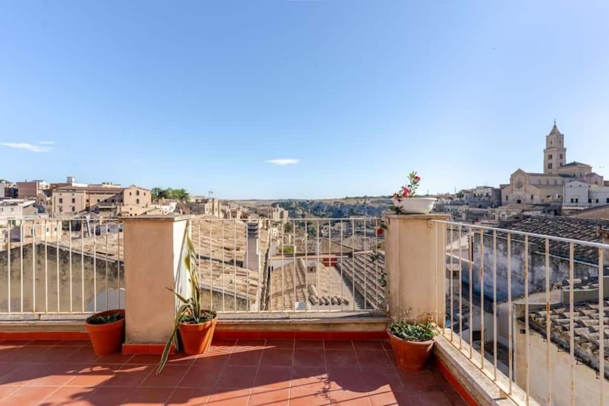 A rooftop terrace with terracotta tiles, potted plants, and a view of historic buildings and a church tower.