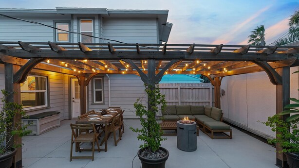 Eine überdachte Terrasse mit einer Holz-Pergola, Lichterketten, einem Esstisch mit Stühlen, einer Feuerstelle und einer Couch.