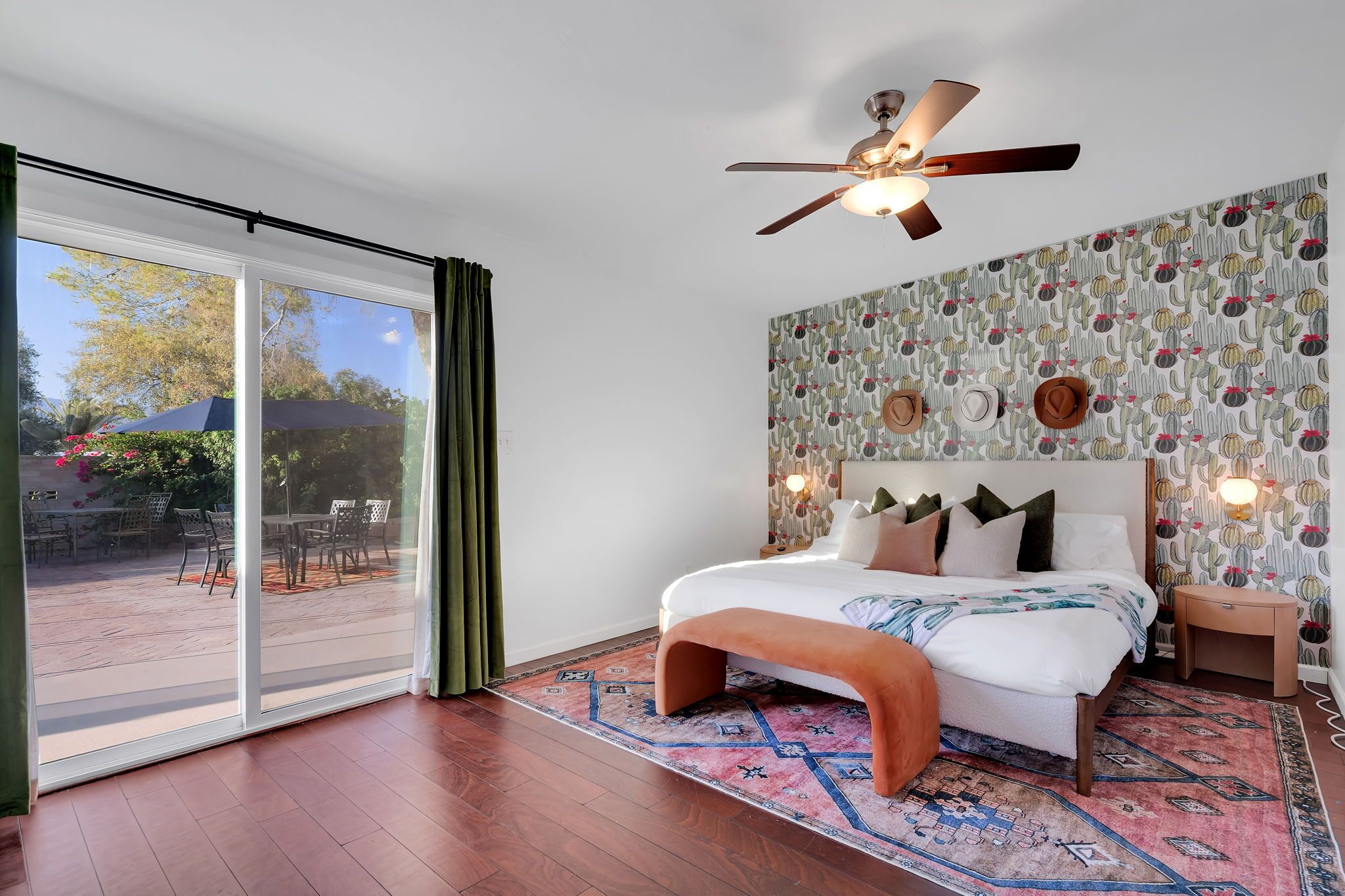 Une chambre avec un grand lit, un ventilateur de plafond, un mur décoré de motifs et une vue sur une terrasse extérieure, accessible par une porte coulissante.