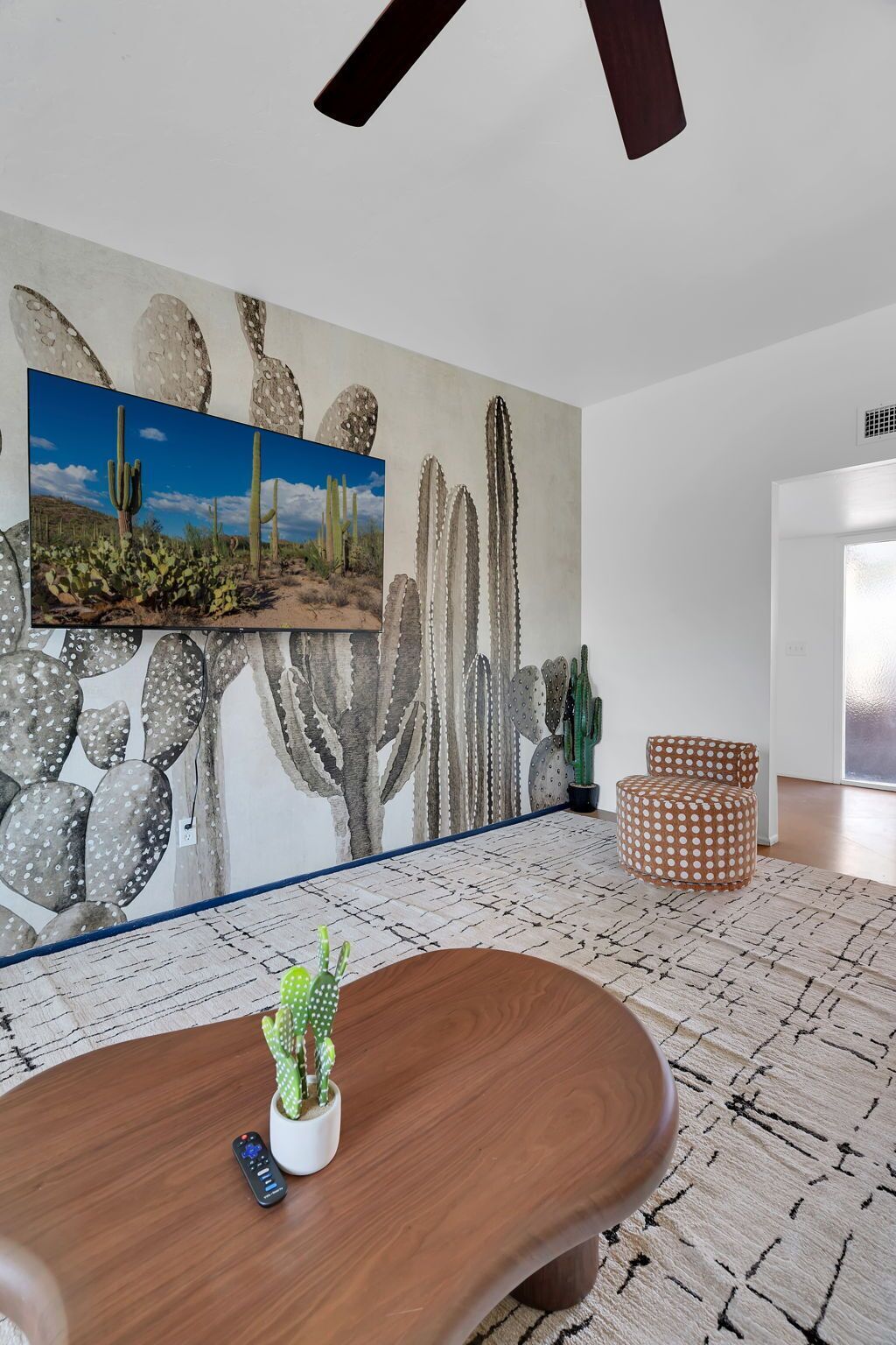 Un salon moderne agrémenté d’une décoration murale sur le thème des cactus, d’une table basse en bois et d’un pouf à pois.