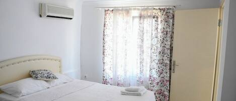 Un dormitorio con cama, una mesita de noche, una ventana con cortinas florales y un equipo de aire acondicionado en la pared.