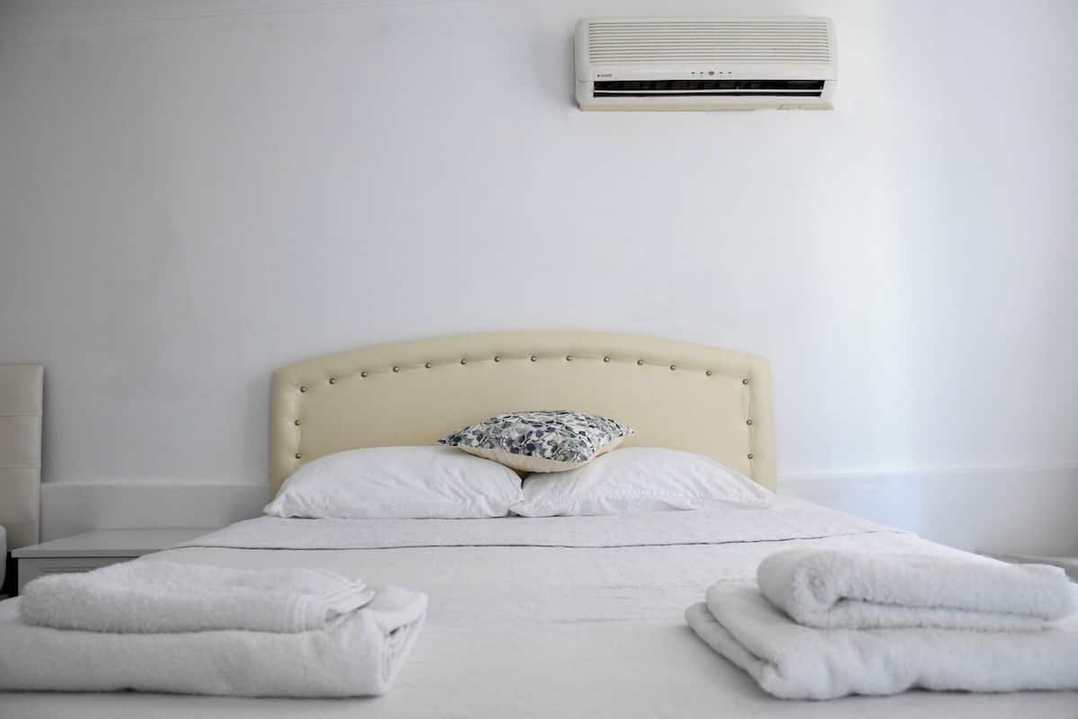 A bed with a beige upholstered headboard, two white pillows, and a floral-patterned pillow. There are two folded white towels on the bed. An air conditioning unit is mounted on the wall above the bed.