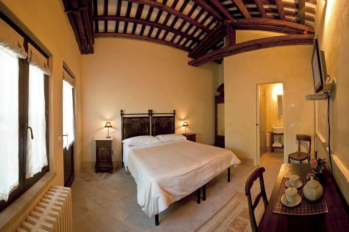 Quarto com uma cama grande, duas luminárias de cabeceira, um painel de cabeceira de madeira, um radiador, uma cadeira e uma mesa pequena com chávenas e um bule.