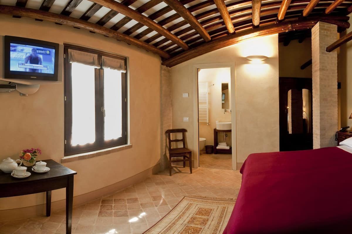 Quarto de hotel com uma cama grande, televisão fixada na parede, uma pequena mesa com bule e flores, e cabeceira de madeira.