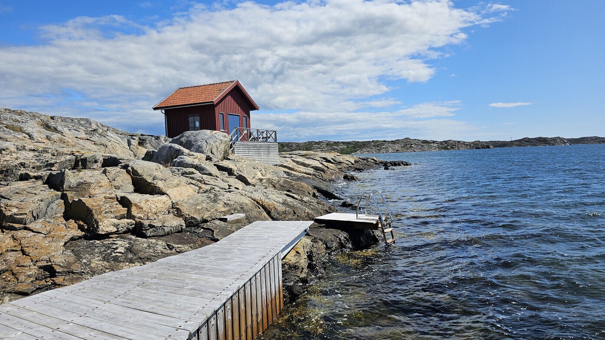 Une côte rocheuse avec une jetée en bois menant à un petit bâtiment rouge doté d’une porte bleue, surplombant une étendue d’eau.