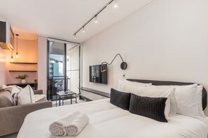 A modern hotel room with a large bed, a sofa, a small table, and a balcony with a view of the city. - Flamingo - Sea Point Lions View (Cape Town)