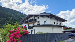 Une maison à deux étages avec un balcon, entourée d’une clôture et d’une végétation luxuriante.