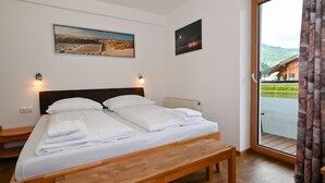 Une chambre à coucher avec un lit en bois, deux tables de chevet, un banc et une grande fenêtre donnant sur un immeuble et des montagnes.