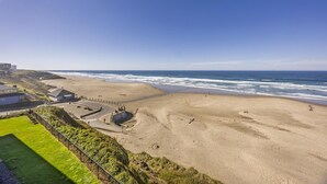 A beach with a curved road leading to a parking area, a grassy area with a fence, and a view of the ocean. - Large Oceanfront Views 2 Kings Bed 2 Bath Lg Spa Tub Romantic Nye Beach Getaway (Newport)