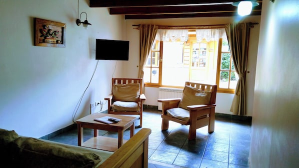 A cozy living room with two wooden chairs, a small table, and a sofa. A flat-screen TV is mounted on the wall, and there's a window with curtains. - Costa Soñada (San Martín de los Andes)