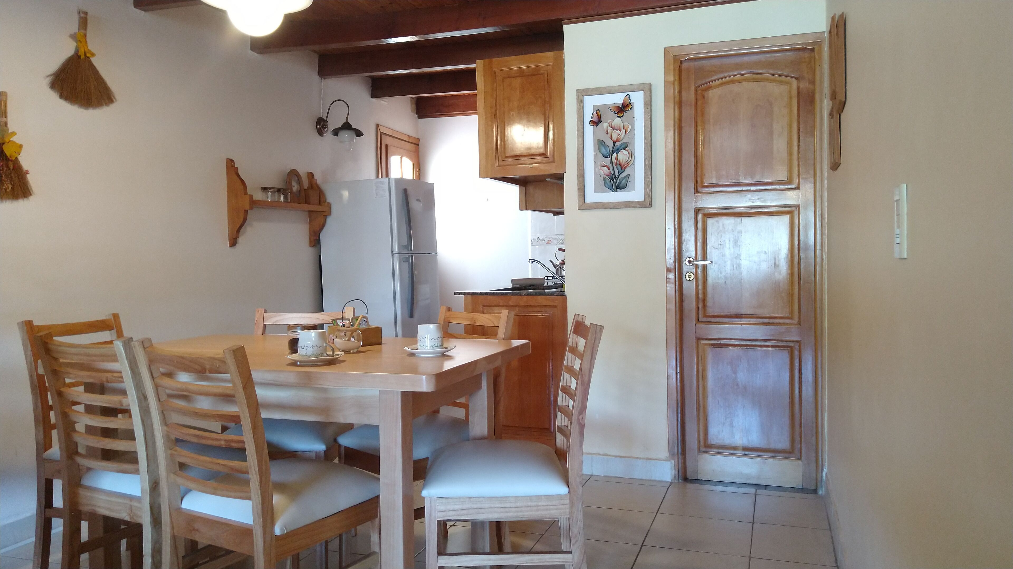 A dining area with a wooden table and chairs, a refrigerator, and a kitchen in the background.