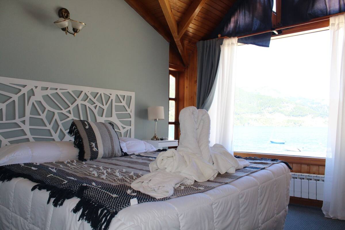 Une chambre avec un grand lit, une tête de lit décorative, une fenêtre donnant sur la mer et une serviette blanche posée sur le lit.