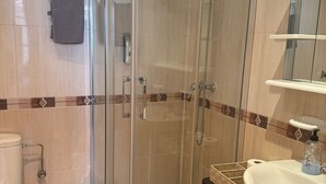 Un baño moderno con ducha envuelta en vidrio, inodoro blanco, lavamanos con dispensador de jabón y un pequeño cubo de basura.