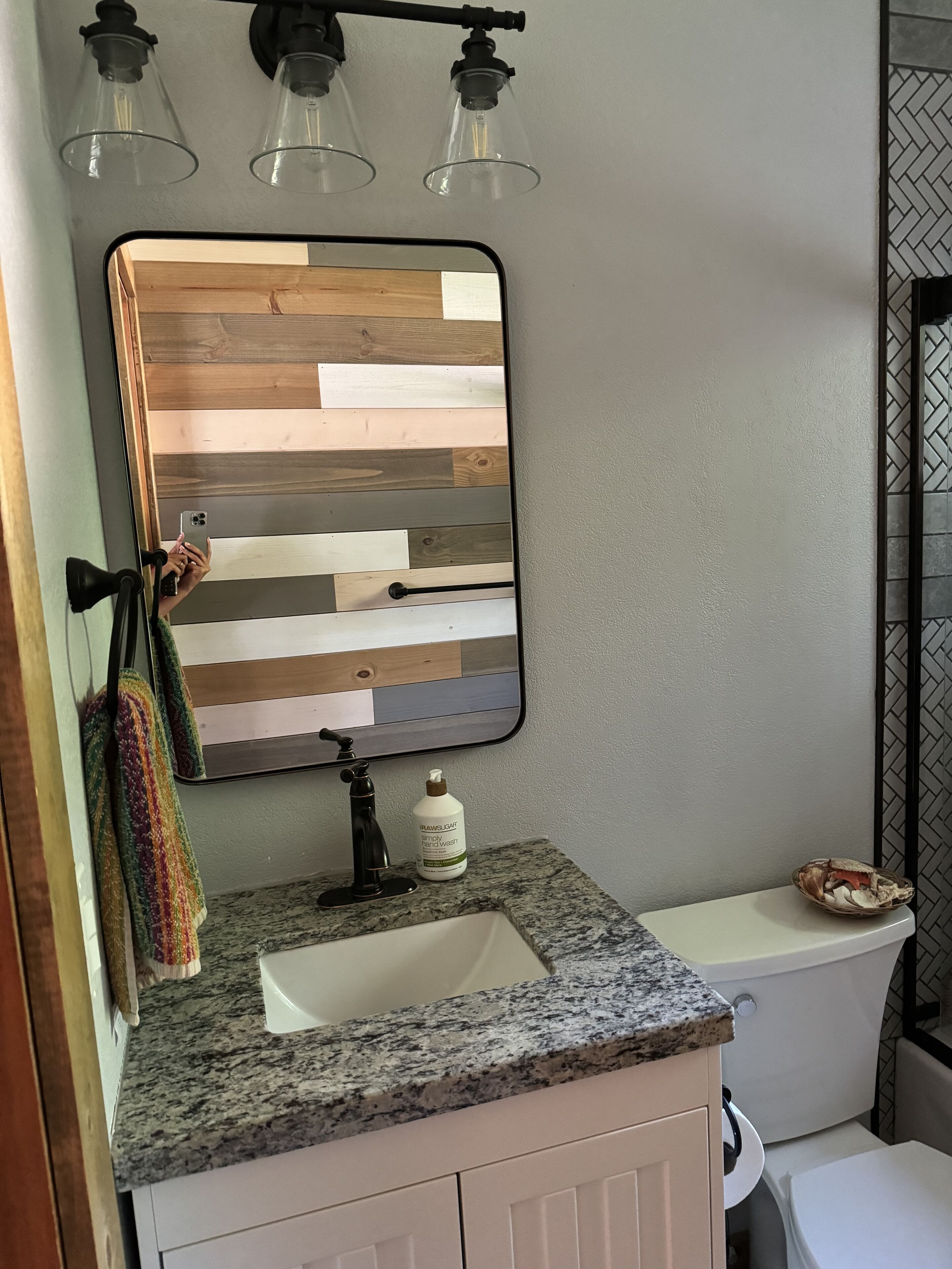 A bathroom with a wooden plank backsplash, a granite countertop, a mirror, a towel rack, a bottle of hand soap, and a toilet.