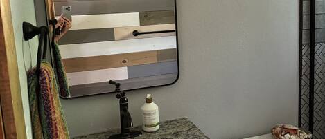 A bathroom with a wooden plank backsplash, a granite countertop, a mirror, a towel rack, a bottle of hand soap, and a toilet.