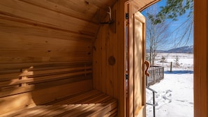 Een houten sauna met een bank, een deur naar buiten en uitzicht op een besneeerd landschap.