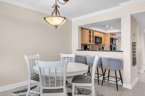 A dining area with a round table and white chairs, a hanging light fixture, and a view into a kitchen with wooden cabinets and modern appliances. - Land's End 11-306 Beach Front - Premier (Treasure Island)