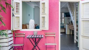 Pared exterior rosa con una pequeña mesa y dos sillas, una ventana con contraventanas y una escalera que lleva a un piso superior.