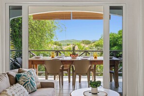 An open-plan living area with a dining table, chairs, and a sofa. A large window offers a view of greenery and hills. - Villá Córal - Villapárk Fóntein (Villapark Fontein)