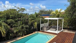 Et rektangulært pool med træterrasse, omgivet af frodig vegetation og en pavillon med siddepladser.