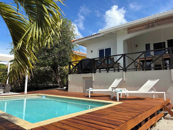 Piscina com deck de madeira, cadeiras de descanso e vista para uma casa com varanda.