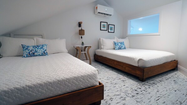 A bedroom with two beds, a small table, a wall-mounted air conditioner, and a window with a view.