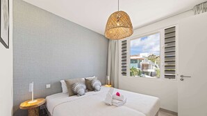 A modern bedroom with a large bed, a bedside table with a lamp, a rattan pendant light, and a view of buildings through a window with blinds. - Palm Paradise-Stylish Island Live-Blue Bay Curaçao (Willemstad)