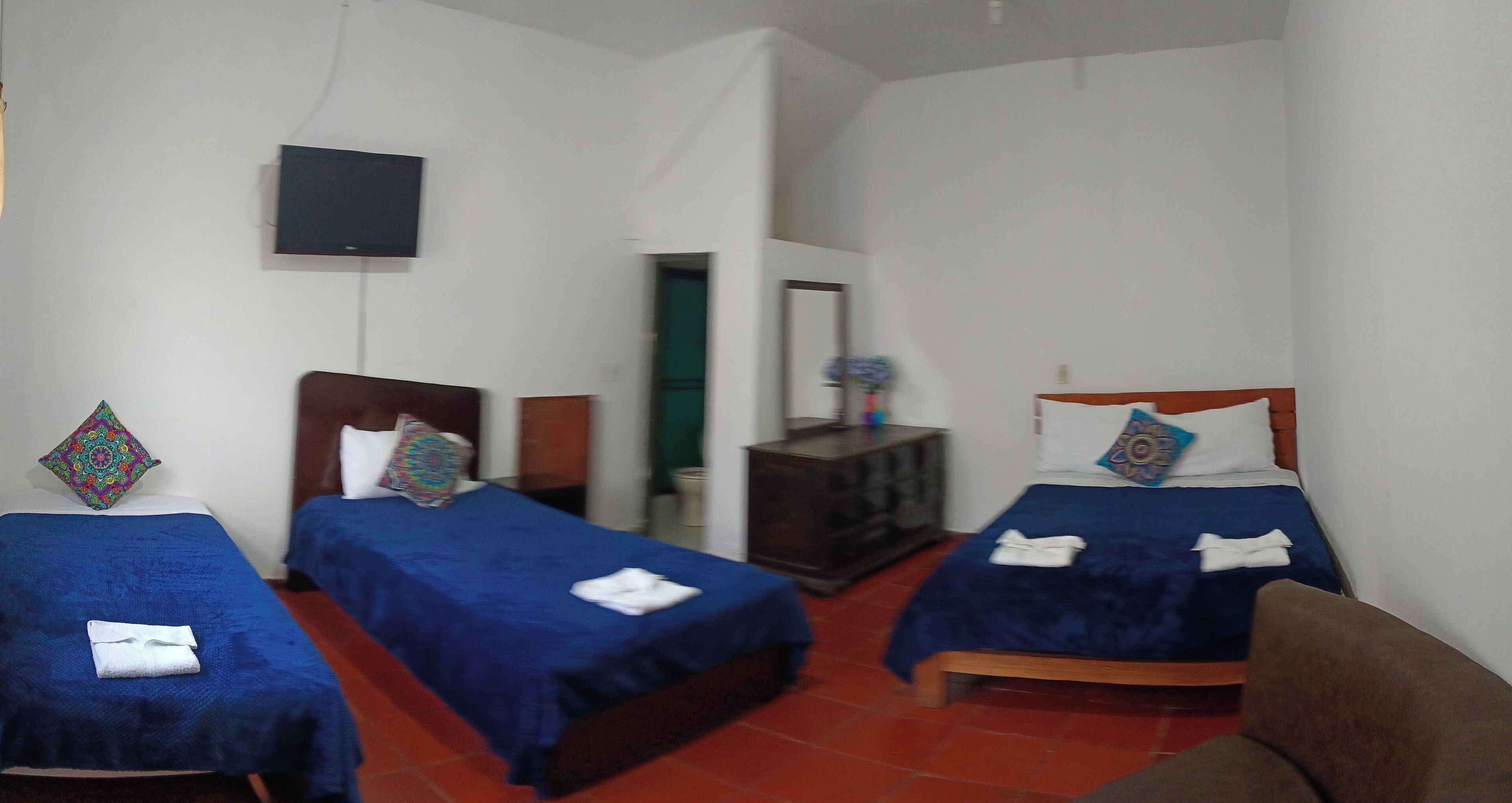 Habitación de hotel con dos camas individuales, cada una con una colcha azul y almohadas decorativas. Se observa un cabecero de madera, un pequeño mueble de madera y un televisor de pantalla plana montado en la pared.