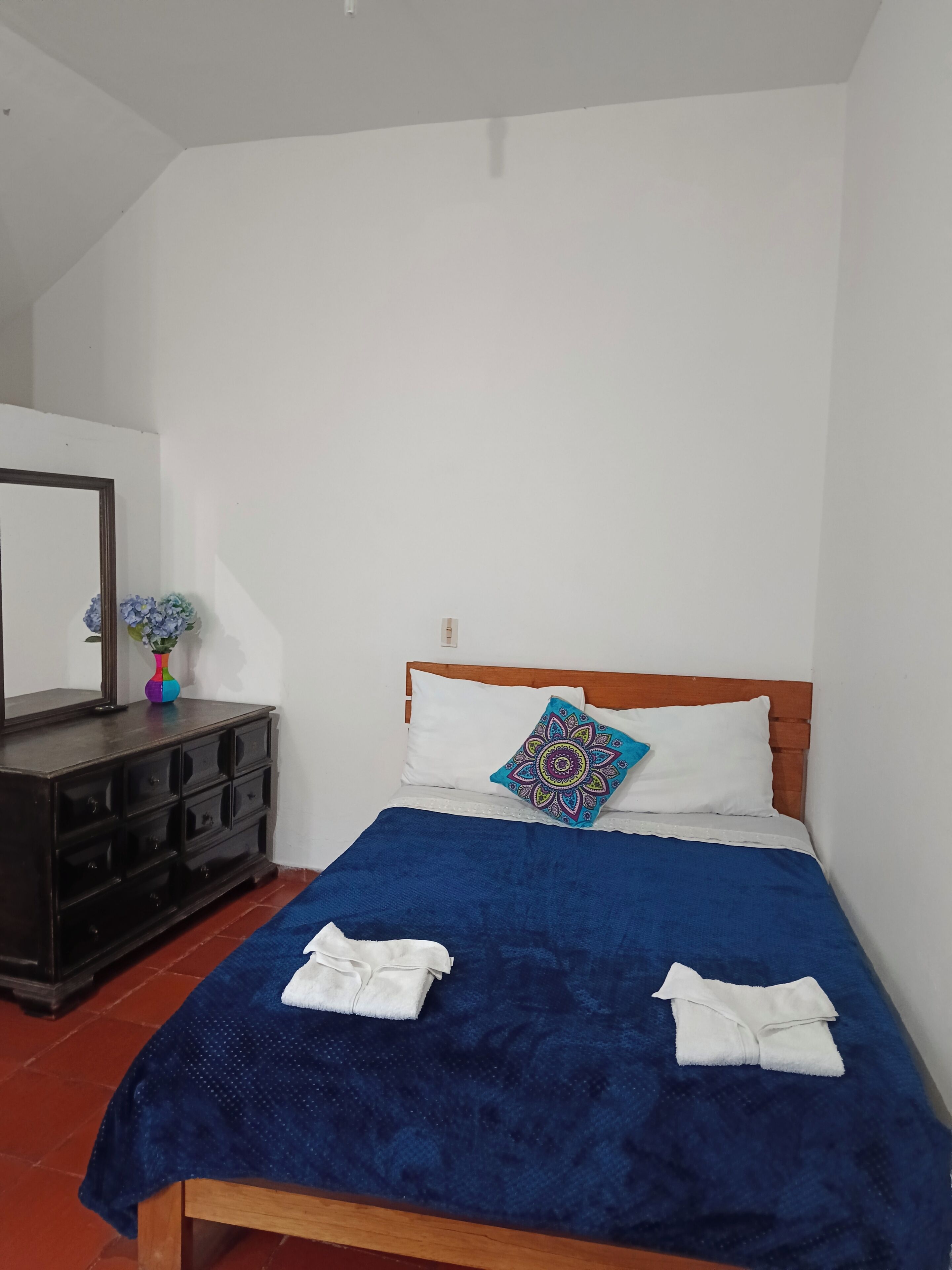 A bedroom with a wooden bed, white pillows, a blue blanket, and a wooden dresser with a vase of flowers.