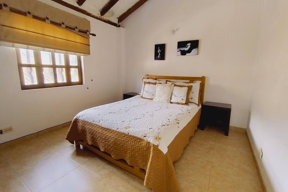 A bedroom with a wooden bed, a window with blinds, two bedside tables, and two wall-mounted pictures.