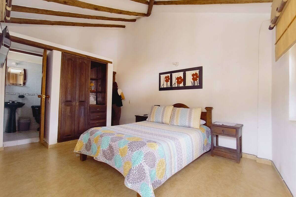 A bedroom with a bed, a nightstand, a wooden wardrobe, a bathroom visible through an open door, and a wall-mounted picture.