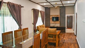 A dining area with a glass table and wooden chairs, a flat-screen TV mounted on a wall, and a ceiling fan.