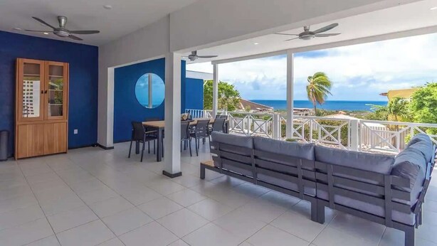 Une chambre spacieuse avec un balcon, un ventilateur de plafond, un grand miroir rond et une vue sur l’océan.