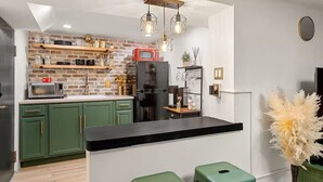 A modern kitchen with green cabinets, a black countertop, and a brick wall. There are two hanging light fixtures, a refrigerator, and a microwave.