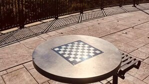 A round table with a chessboard in the foreground, a metal fence, and a city skyline in the background.
