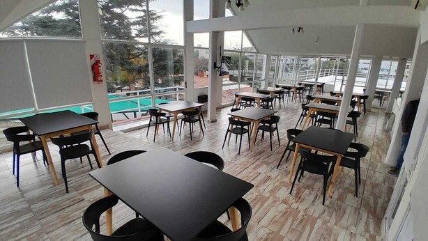 A spacious indoor area with multiple tables and chairs, large windows offering views of the outside, and a high ceiling with hanging lights. - HOTEL CRISTAL (Villa Carlos Paz)