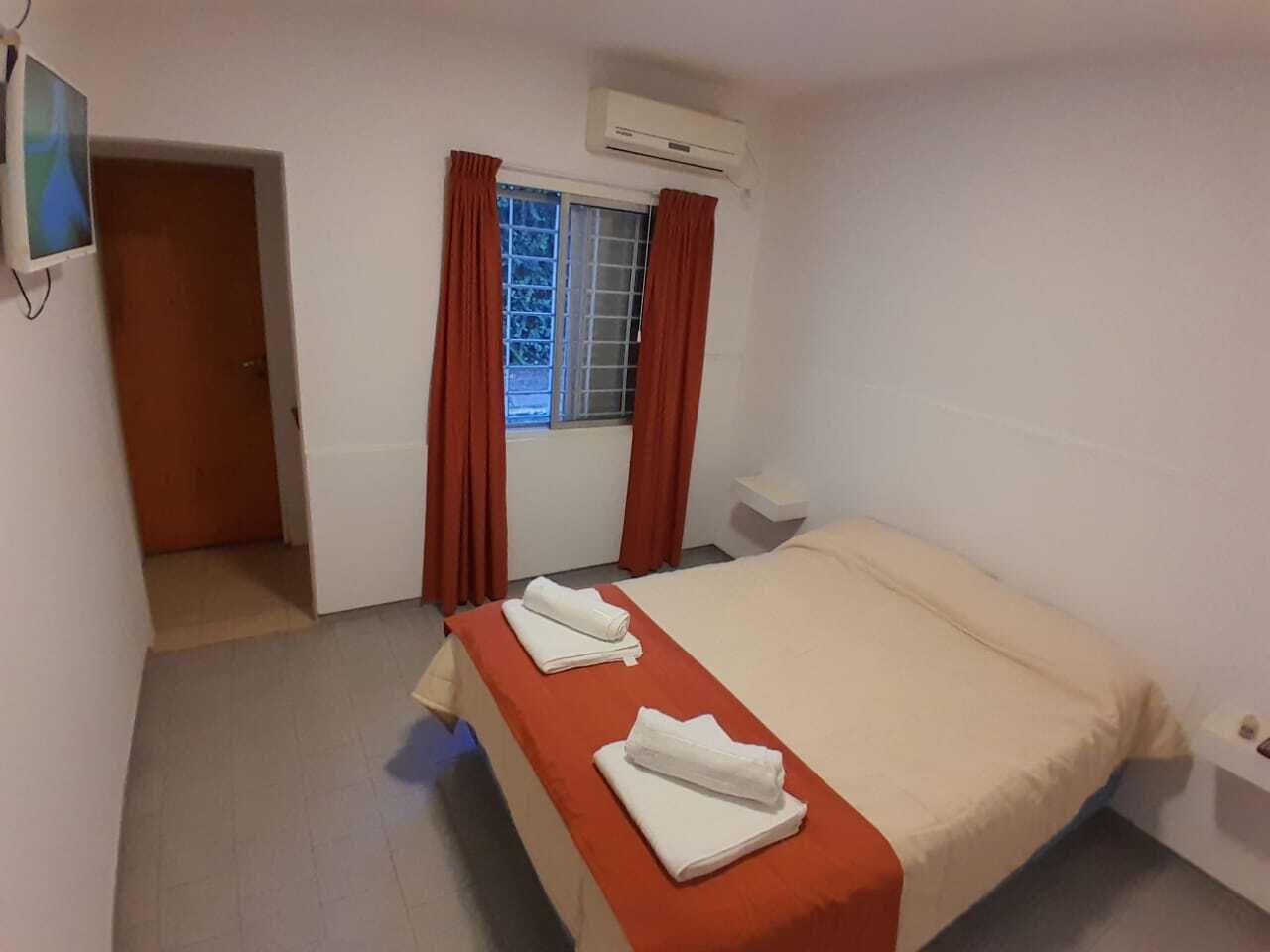 A hotel room with a bed, a window with curtains, an air conditioning unit, and a television mounted on the wall.